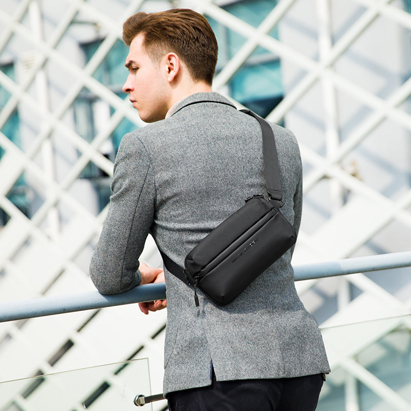 Man wearing black waterproof crossbody bag with multiple compartments over gray blazer, modern architectural backdrop | Mark Ryden Global