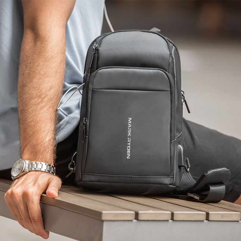 Dark gray Mark Ryden Mini Compacto lightweight minimalist tablet sling bag with multi-layer compartments, resting on a wooden bench.