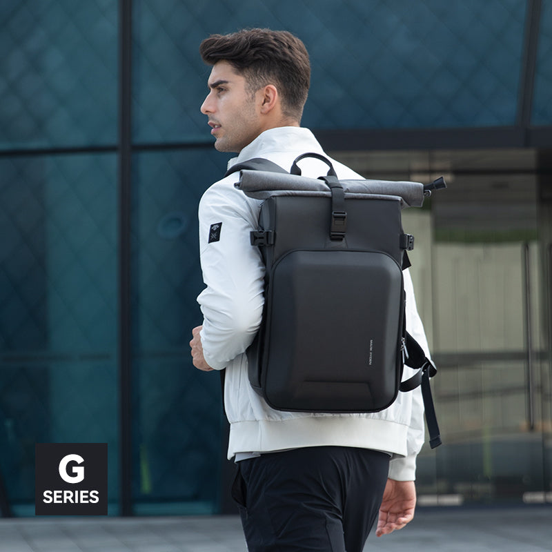 Black large-capacity camera backpack with roll-top closure, worn by a man in a white jacket and black pants, against a modern glass building background, Mark Ryden Global G Series