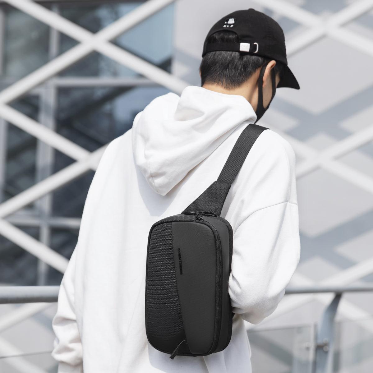 Stylish waterproof black crossbody bag with multiple compartments worn by man in white hoodie near modern building