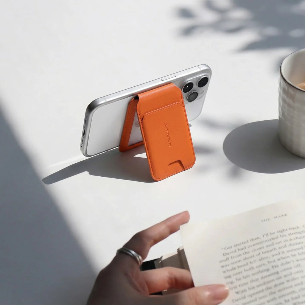 MagFold orange slim MagSafe foldable card holder wallet attached to silver iPhone, functioning as a phone stand with a book and cup nearby.