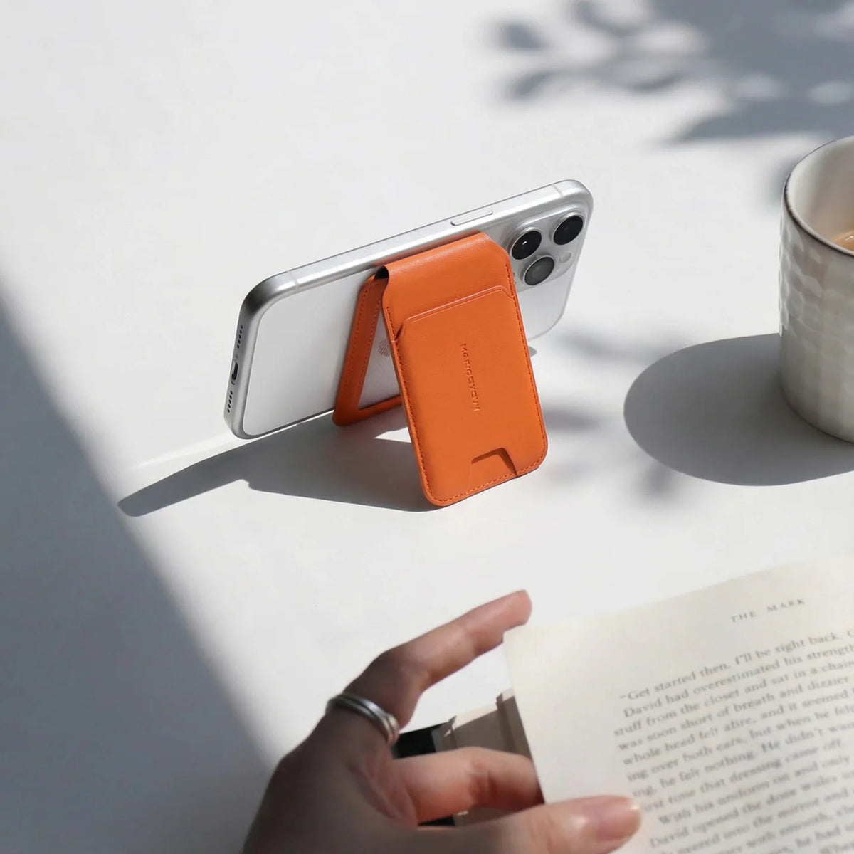 MagFold orange slim MagSafe foldable card holder wallet attached to silver iPhone, functioning as a phone stand with a book and cup nearby.