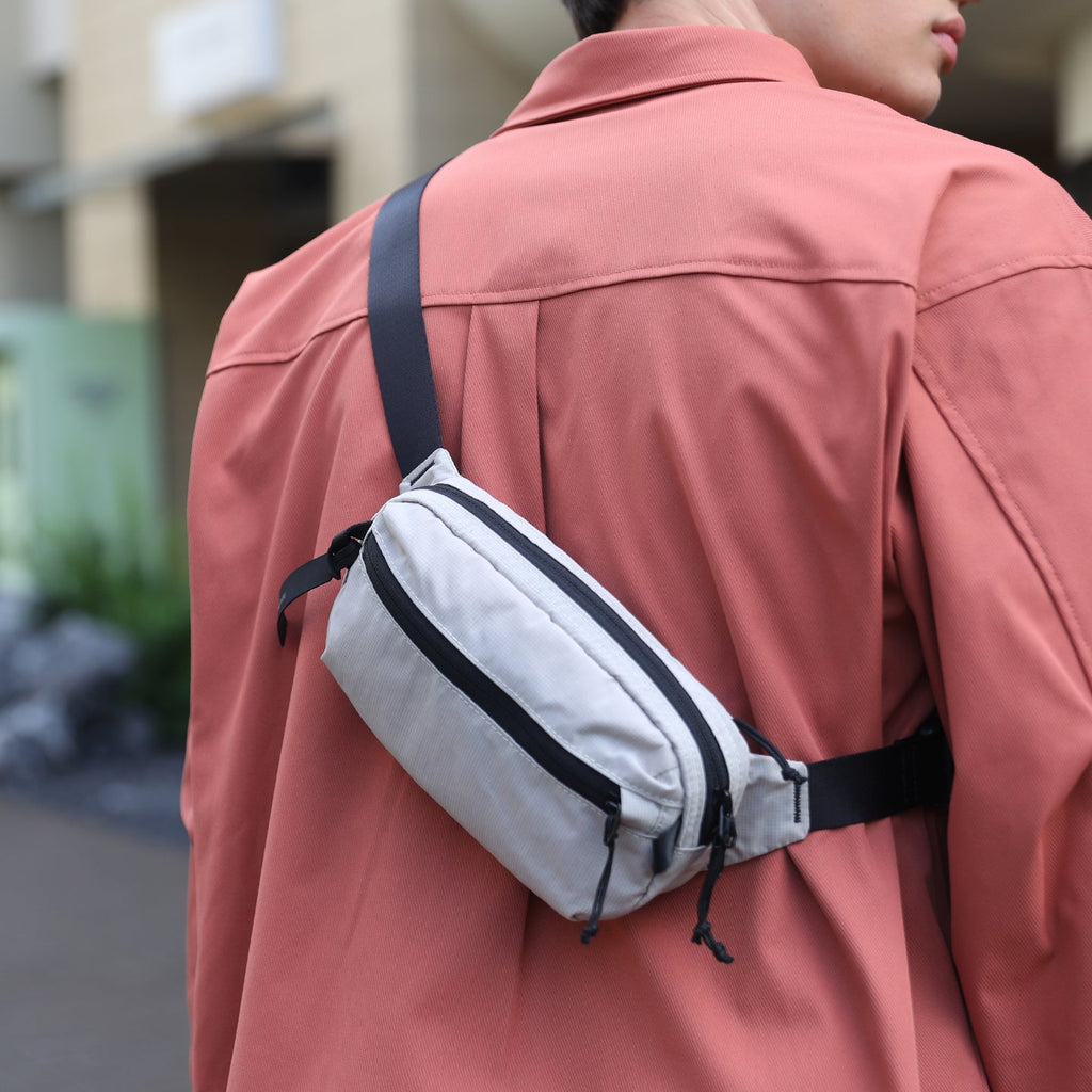 Light gray AeroSling ultra-light anti-theft crossbody bag with RFID-safe, black zippers and waist strap, worn across the back by a person in a coral jacket.