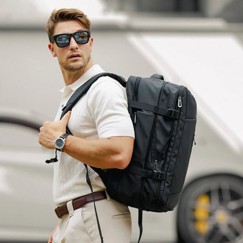 Black MARK RYDEN Expandos Max anti-theft compression backpack with multiple zippers, adjustable straps, and a spacious design, worn by a man in a white polo and beige pants for travel.